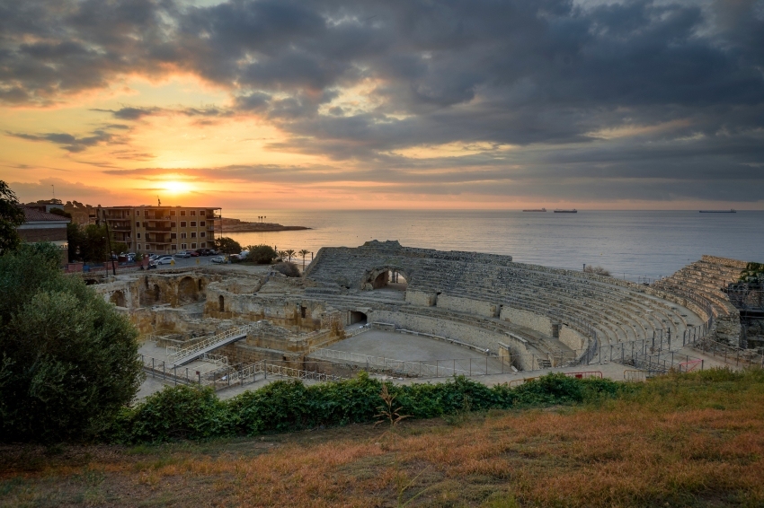 Ruta por la historia romana de Tarragona: visita guiada al anfiteatro, circo y murallas, con teatralización nocturna en verano Descubrir Tarragona a través de una ruta romana es sumergirse en 2 000 años de historia viva, recorriendo monumentos declarados Patrimonio de la Humanidad por la Unesco. La experiencia empieza junto a las imponentes murallas, que protegían la antigua Tarraco y hoy sirven de puerta de entrada a un viaje único en el tiempo. Un guía especializado va desgranando los secretos, leyendas y anécdotas de la ciudad mientras se pasea por el casco antiguo, admirando la catedral y los restos arqueológicos que conectan pasado y presente. El recorrido atraviesa el Foro provincial y el Circo romano, donde el bullicio de las carreras de caballos y aurigas aún resuena en las bóvedas y gradas originales. La subida a la Torre del Pretorio regala vistas espectaculares sobre las azoteas históricas y el Mediterráneo. El broche final lo pone el Anfiteatro, excavado en roca junto al mar, escenario de luchas de gladiadores y grandes espectáculos, cuya estructura sorprende por su majestuosidad y conservación. En verano, la visita se transforma en una auténtica teatralización nocturna. El guía, vestido como ciudadano romano, interpreta la historia con pasión, haciendo revivir personajes y escenas antiguas bajo una iluminación evocadora. Paneles con recreaciones digitales permiten imaginar el esplendor monumental de Tarraco, mientras el paseo nocturno convierte murallas y plazas en un verdadero teatro arqueológico. “Las Noches de Tarraco” son una experiencia sensorial y educativa, perfecta para familias, grupos de amigos y viajeros curiosos, que buscan algo más que una simple visita turística. Adentrarse en la Tarragona romana es caminar entre leyendas, recorrer plazas donde antes se celebraban carreras y luchas, y dejarse sorprender por la fuerza de una ciudad que sigue viva en sus piedras, sus vistas y sus historias. Una oportunidad para vivir la historia en primera persona, aprendiendo y disfrutando de la magia de la noche, el mar y el legado de Roma en Tarragona. ruta-de-tarraco-empremta-de-roma