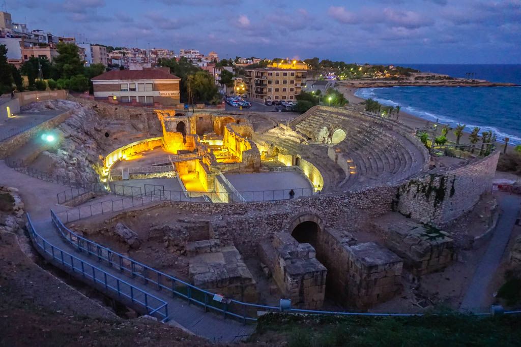 tarragonaromana28Ruta por la historia romana de Tarragona: visita guiada al anfiteatro, circo y murallas, con teatralización nocturna en verano Descubrir Tarragona a través de una ruta romana es sumergirse en 2 000 años de historia viva, recorriendo monumentos declarados Patrimonio de la Humanidad por la Unesco. La experiencia empieza junto a las imponentes murallas, que protegían la antigua Tarraco y hoy sirven de puerta de entrada a un viaje único en el tiempo. Un guía especializado va desgranando los secretos, leyendas y anécdotas de la ciudad mientras se pasea por el casco antiguo, admirando la catedral y los restos arqueológicos que conectan pasado y presente. El recorrido atraviesa el Foro provincial y el Circo romano, donde el bullicio de las carreras de caballos y aurigas aún resuena en las bóvedas y gradas originales. La subida a la Torre del Pretorio regala vistas espectaculares sobre las azoteas históricas y el Mediterráneo. El broche final lo pone el Anfiteatro, excavado en roca junto al mar, escenario de luchas de gladiadores y grandes espectáculos, cuya estructura sorprende por su majestuosidad y conservación. En verano, la visita se transforma en una auténtica teatralización nocturna. El guía, vestido como ciudadano romano, interpreta la historia con pasión, haciendo revivir personajes y escenas antiguas bajo una iluminación evocadora. Paneles con recreaciones digitales permiten imaginar el esplendor monumental de Tarraco, mientras el paseo nocturno convierte murallas y plazas en un verdadero teatro arqueológico. “Las Noches de Tarraco” son una experiencia sensorial y educativa, perfecta para familias, grupos de amigos y viajeros curiosos, que buscan algo más que una simple visita turística. Adentrarse en la Tarragona romana es caminar entre leyendas, recorrer plazas donde antes se celebraban carreras y luchas, y dejarse sorprender por la fuerza de una ciudad que sigue viva en sus piedras, sus vistas y sus historias. Una oportunidad para vivir la historia en primera persona, aprendiendo y disfrutando de la magia de la noche, el mar y el legado de Roma en Tarragona.